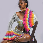 A model wearing a multicolored beaded crochet skirt and top set with long sleeves and a V-neckline, seated against a plain background.