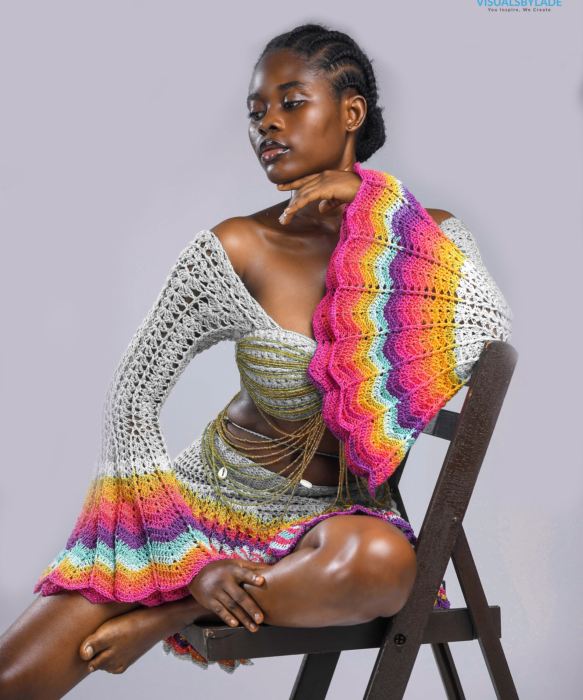 A model wearing a multicolored beaded crochet skirt and top set with long sleeves and a V-neckline, seated against a plain background.
