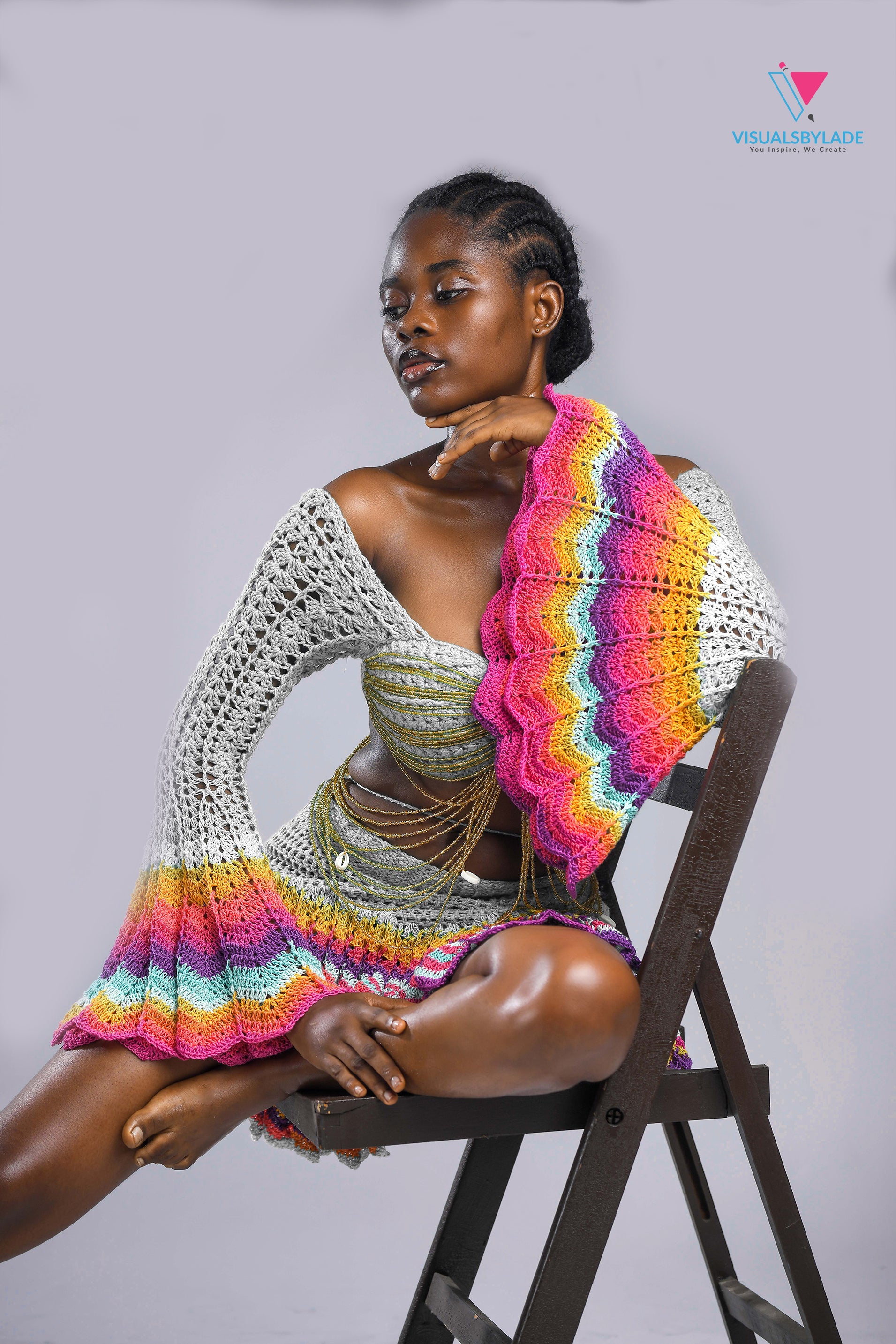 A model wearing a multicolored beaded crochet skirt and top set with long sleeves and a V-neckline, seated against a plain background.
